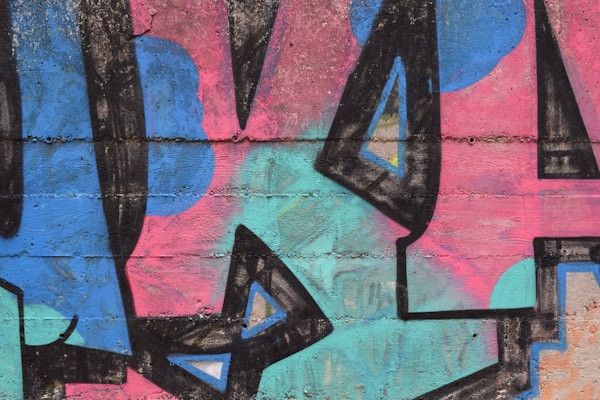 A vibrant graffiti mural featuring a combination of angular and irregular shapes, rendered in bold colors on a textured concrete wall. The artwork includes large abstract forms in shades of blue, pink, and turquoise, with thick black outlines and occasional triangular embellishments.