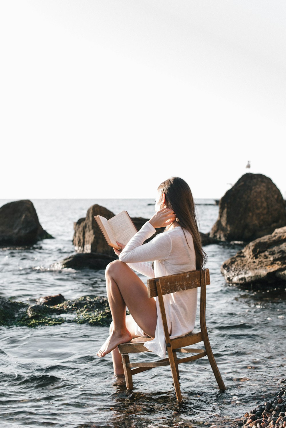 donna in abito bianco che si siede sulla sedia di legno marrone che legge il libro sulla spiaggia durante il giorno