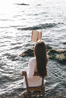 woman in white long sleeve shirt sitting on chair reading book on sea shore during daytime