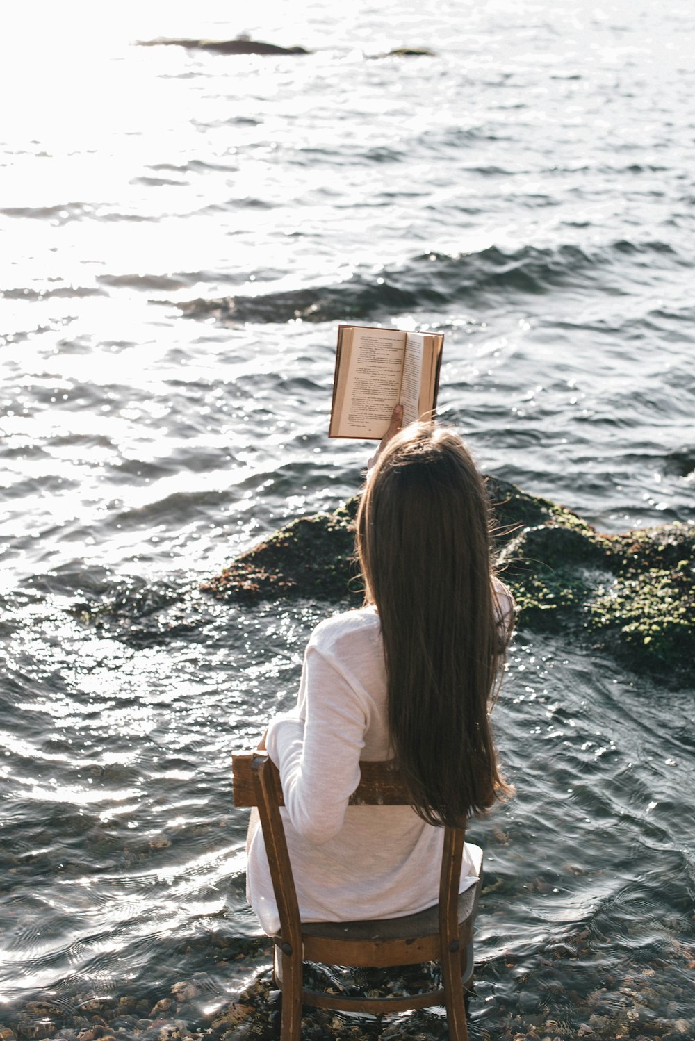 woman in white long sleeve shirt sitting on chair reading book on sea shore during daytime