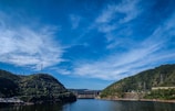 A scenic landscape with a wide river flanked by two green hills under a clear blue sky. A dam stretches across the river in the background, and electrical towers can be seen on the hills.