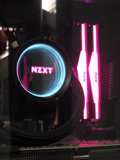 A close-up view of a computer's internal components featuring a liquid cooling system from NZXT with an illuminated circular display in blue and pink hues. Adjacent are two sticks of XPG RAM that glow brightly in pink.