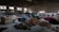 A spacious warehouse filled with large piles of various colored fabric scattered across the floor. There are stacks of filled bags in the background, with some metal shelving units. Two people are walking near the piles, suggesting activity related to sorting or organizing the textiles. The lighting in the space is dim, with some natural light filtering in through the large windows.