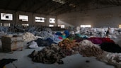 A spacious warehouse filled with large piles of various colored fabric scattered across the floor. There are stacks of filled bags in the background, with some metal shelving units. Two people are walking near the piles, suggesting activity related to sorting or organizing the textiles. The lighting in the space is dim, with some natural light filtering in through the large windows.