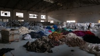 A spacious warehouse filled with large piles of various colored fabric scattered across the floor. There are stacks of filled bags in the background, with some metal shelving units. Two people are walking near the piles, suggesting activity related to sorting or organizing the textiles. The lighting in the space is dim, with some natural light filtering in through the large windows.