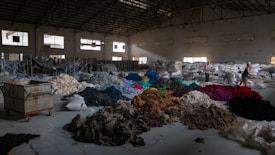 A spacious warehouse filled with large piles of various colored fabric scattered across the floor. There are stacks of filled bags in the background, with some metal shelving units. Two people are walking near the piles, suggesting activity related to sorting or organizing the textiles. The lighting in the space is dim, with some natural light filtering in through the large windows.