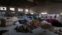 A spacious warehouse filled with large piles of various colored fabric scattered across the floor. There are stacks of filled bags in the background, with some metal shelving units. Two people are walking near the piles, suggesting activity related to sorting or organizing the textiles. The lighting in the space is dim, with some natural light filtering in through the large windows.