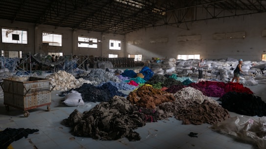 A spacious warehouse filled with large piles of various colored fabric scattered across the floor. There are stacks of filled bags in the background, with some metal shelving units. Two people are walking near the piles, suggesting activity related to sorting or organizing the textiles. The lighting in the space is dim, with some natural light filtering in through the large windows.