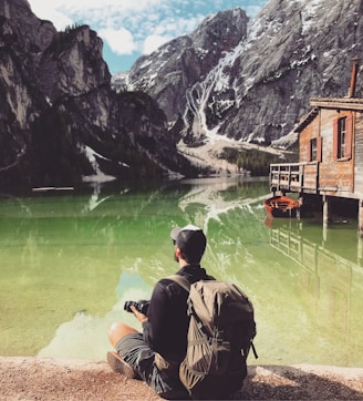 man in brown jacket sitting on brown rock near body of water during daytime