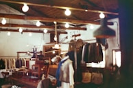 A rustic clothing store interior with hanging light bulbs illuminating the space. Wooden beams and racks display various garments, including coats and jackets. Wooden furniture and vintage decor enhance the cozy ambiance.