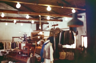 A rustic clothing store interior with hanging light bulbs illuminating the space. Wooden beams and racks display various garments, including coats and jackets. Wooden furniture and vintage decor enhance the cozy ambiance.