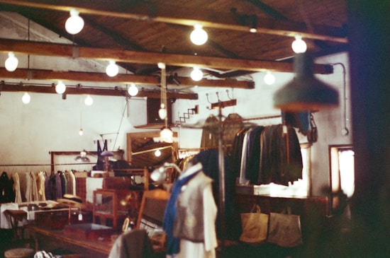 A rustic clothing store interior with hanging light bulbs illuminating the space. Wooden beams and racks display various garments, including coats and jackets. Wooden furniture and vintage decor enhance the cozy ambiance.