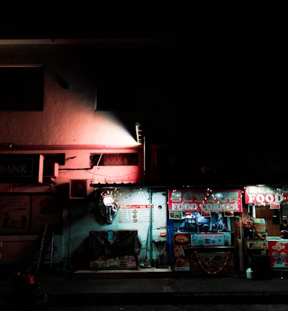 A dimly lit street scene with a small food stall named 'Food Corner'. The stall is adorned with colorful signs and decorations, illuminated by soft lights. The surrounding area is dark, highlighting the bright storefront. A person can be seen partially behind the counter, and there are various items like a menu board and food posters visible.