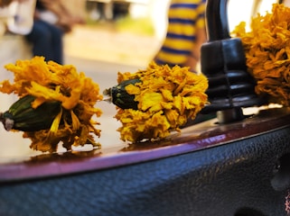 A close-up of dried yellow marigold flowers strung together and wrapped around a metallic object, possibly a motorcycle handlebar. In the background, there is an out-of-focus person wearing a blue and yellow striped shirt.