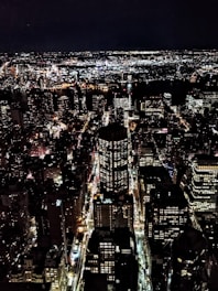 A sprawling cityscape at night with brightly lit skyscrapers and streets. The city is alive with clusters of lights from buildings, streetlights, and traffic creating a dynamic and vibrant urban environment. The sky above is dark, contrasting with the intense glow of the city.
