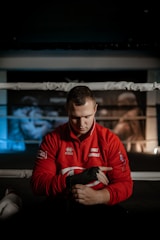 A person wearing a red jacket sits in a boxing ring, wrapping their hands with black hand wraps. A focused expression is visible as they prepare for training or a match. Boxing posters can be seen in the blurred background, adding to the athletic environment.
