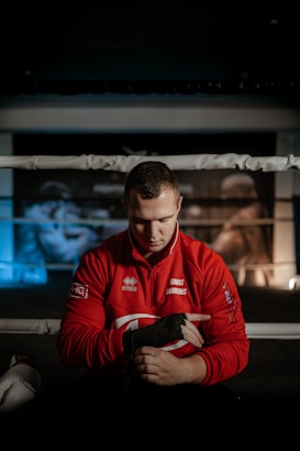 A person wearing a red jacket sits in a boxing ring, wrapping their hands with black hand wraps. A focused expression is visible as they prepare for training or a match. Boxing posters can be seen in the blurred background, adding to the athletic environment.
