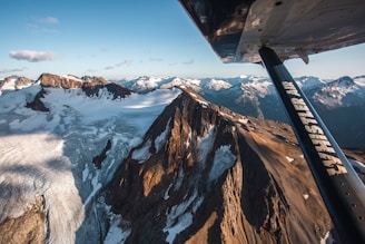 A breathtaking aerial view of rugged mountain peaks and expansive glaciers, taken from an airplane. The mountains are rugged with snow patches, and the light creates dramatic shadows. The airplane wing, marked with 'Whistler Air,' is prominently visible in the foreground, adding depth to the scene.