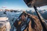 A breathtaking aerial view of rugged mountain peaks and expansive glaciers, taken from an airplane. The mountains are rugged with snow patches, and the light creates dramatic shadows. The airplane wing, marked with 'Whistler Air,' is prominently visible in the foreground, adding depth to the scene.