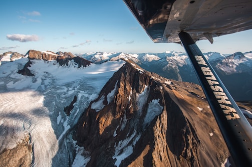 A breathtaking aerial view of rugged mountain peaks and expansive glaciers, taken from an airplane. The mountains are rugged with snow patches, and the light creates dramatic shadows. The airplane wing, marked with 'Whistler Air,' is prominently visible in the foreground, adding depth to the scene.
