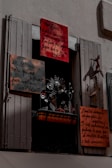 A window display featuring several hand-painted signs with French text. The signs vary in size and are attached to a window frame with shutters partially open. Below the signs, there is a decorative metal sculpture with floral and bird motifs. The overall scene is shadowed, giving a slightly mysterious atmosphere.