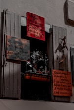 A window display featuring several hand-painted signs with French text. The signs vary in size and are attached to a window frame with shutters partially open. Below the signs, there is a decorative metal sculpture with floral and bird motifs. The overall scene is shadowed, giving a slightly mysterious atmosphere.