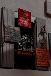 A window display featuring several hand-painted signs with French text. The signs vary in size and are attached to a window frame with shutters partially open. Below the signs, there is a decorative metal sculpture with floral and bird motifs. The overall scene is shadowed, giving a slightly mysterious atmosphere.