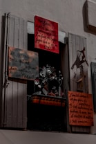 A window display featuring several hand-painted signs with French text. The signs vary in size and are attached to a window frame with shutters partially open. Below the signs, there is a decorative metal sculpture with floral and bird motifs. The overall scene is shadowed, giving a slightly mysterious atmosphere.