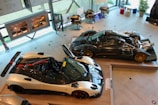 An exhibition space featuring high-performance sports cars. Two sleek racing cars are displayed prominently, with one showcasing an exposed engine and intricate mechanical components. The brightly lit room has large windows and several framed diagrams on the walls. Engine models and car parts are neatly arranged on stands.