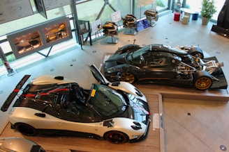 An exhibition space featuring high-performance sports cars. Two sleek racing cars are displayed prominently, with one showcasing an exposed engine and intricate mechanical components. The brightly lit room has large windows and several framed diagrams on the walls. Engine models and car parts are neatly arranged on stands.
