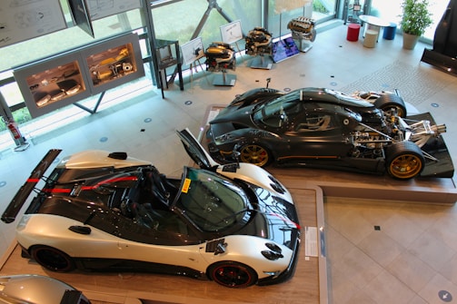 An exhibition space featuring high-performance sports cars. Two sleek racing cars are displayed prominently, with one showcasing an exposed engine and intricate mechanical components. The brightly lit room has large windows and several framed diagrams on the walls. Engine models and car parts are neatly arranged on stands.