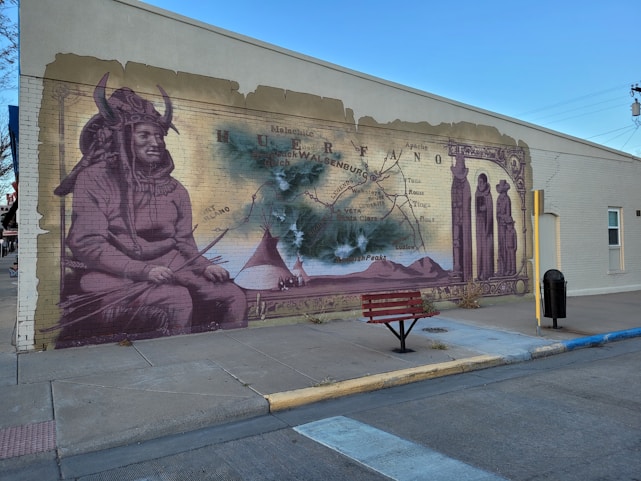 A mural painted on the side of a building depicts indigenous imagery, featuring a person wearing traditional clothing and a horned headdress. There is a vintage map in the background with geographic markers, along with tepees and mountains in the central part of the mural. The artwork incorporates sepia tones and appears to be weathered as if blending historical elements with cultural significance. In front of the mural is a red bench on the sidewalk.