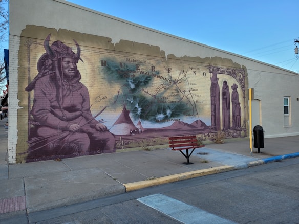 A mural painted on the side of a building depicts indigenous imagery, featuring a person wearing traditional clothing and a horned headdress. There is a vintage map in the background with geographic markers, along with tepees and mountains in the central part of the mural. The artwork incorporates sepia tones and appears to be weathered as if blending historical elements with cultural significance. In front of the mural is a red bench on the sidewalk.