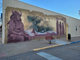A mural painted on the side of a building depicts indigenous imagery, featuring a person wearing traditional clothing and a horned headdress. There is a vintage map in the background with geographic markers, along with tepees and mountains in the central part of the mural. The artwork incorporates sepia tones and appears to be weathered as if blending historical elements with cultural significance. In front of the mural is a red bench on the sidewalk.