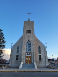 A large, historic church building with a central steeple topped by a cross. The facade features tall, narrow arched windows and a prominent wooden door with a small staircase leading up to it. The structure appears to be made from stone or concrete. The sky is clear and blue, with a few clouds visible in the background. Adjacent to the church, trees and some residential buildings are visible.