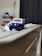 A small toy model of a blue and white vehicle is placed on a narrow edge. The background is blurred, featuring what looks like a dark patterned curtain and some indistinct objects.
