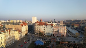 A cityscape featuring historic European architecture with ornate facades and red-tiled roofs, set against a backdrop of modern buildings. A river flows through the right side, bordered by trees and parked cars. The streets below are bustling with vehicles and pedestrians.