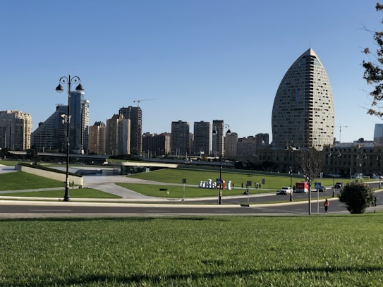 A cityscape featuring a park in the foreground, characterized by lush green lawns and pathways. Tall, modern buildings with diverse architectural designs form the backdrop. Among them, a uniquely shaped oval building stands out. The sky is clear and blue, giving the scene a bright and open feel.