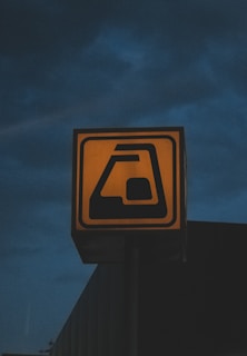 A square, illuminated sign stands against a dark, cloudy sky. The sign features a stylized image that appears to represent an ATM or cash machine, with a machine-like shape holding a card.