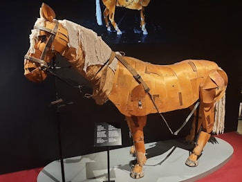 A mechanical horse structure composed of wooden panels and fabric, giving it the appearance of a worn-out theatrical or artistic prop. It stands on a display platform, with a visible plaque providing information. The model is detailed, including metal reinforcements and worn fabric strands that simulate a mane.