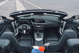 The interior of a modern convertible car with a sleek, black leather finish and a sophisticated dashboard featuring a digital display and various controls. The steering wheel prominently displays the BMW logo. Two plush front seats are visible with a colorful soft toy placed between them. The background shows an empty road and rails.