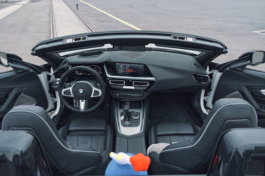 The interior of a modern convertible car with a sleek, black leather finish and a sophisticated dashboard featuring a digital display and various controls. The steering wheel prominently displays the BMW logo. Two plush front seats are visible with a colorful soft toy placed between them. The background shows an empty road and rails.