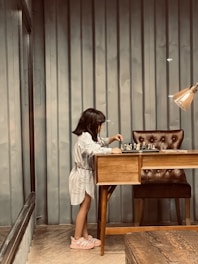 A young girl with shoulder-length hair, wearing a light-colored dress and pink shoes, is standing at a wooden desk. She is engaged in playing chess, with a well-organized chessboard in front of her. The room has a modern industrial feel, featuring corrugated metal walls, a cushioned chair, and an overhead lamp providing warm lighting.