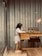 A young girl with shoulder-length hair, wearing a light-colored dress and pink shoes, is standing at a wooden desk. She is engaged in playing chess, with a well-organized chessboard in front of her. The room has a modern industrial feel, featuring corrugated metal walls, a cushioned chair, and an overhead lamp providing warm lighting.