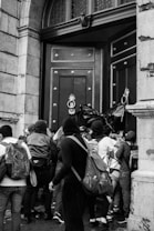 A group of people wearing backpacks and hooded clothing gather closely outside a large, intricately designed double door. The setting appears to be the entryway of a stone building with an arched top. Some individuals are actively attempting to push the door open.