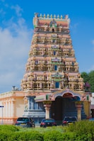 A vibrant and intricately detailed Hindu temple with a tall, ornate gopuram adorned with various colorful figures and patterns. The temple is surrounded by lush greenery, and several cars are parked in front of it. A clear blue sky serves as the backdrop.