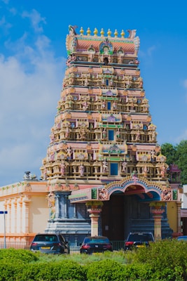 A vibrant and intricately detailed Hindu temple with a tall, ornate gopuram adorned with various colorful figures and patterns. The temple is surrounded by lush greenery, and several cars are parked in front of it. A clear blue sky serves as the backdrop.