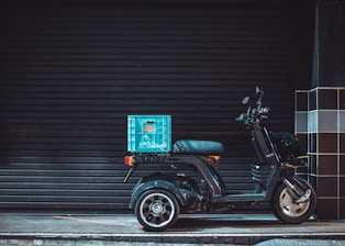 A three-wheeled black scooter parked on a dimly lit street with a blue crate attached to its rear. The background features a black, closed shutter and the sidewalk has a grid-patterned black and white tiled wall nearby.