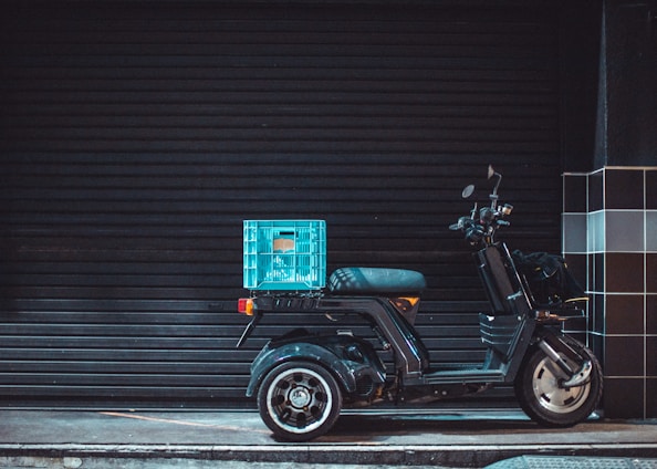 A three-wheeled black scooter parked on a dimly lit street with a blue crate attached to its rear. The background features a black, closed shutter and the sidewalk has a grid-patterned black and white tiled wall nearby.