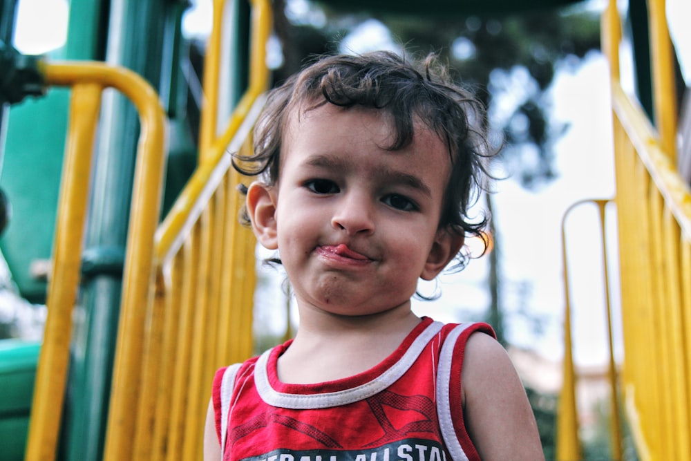 girl in red tank top standing near yellow metal fence during daytime